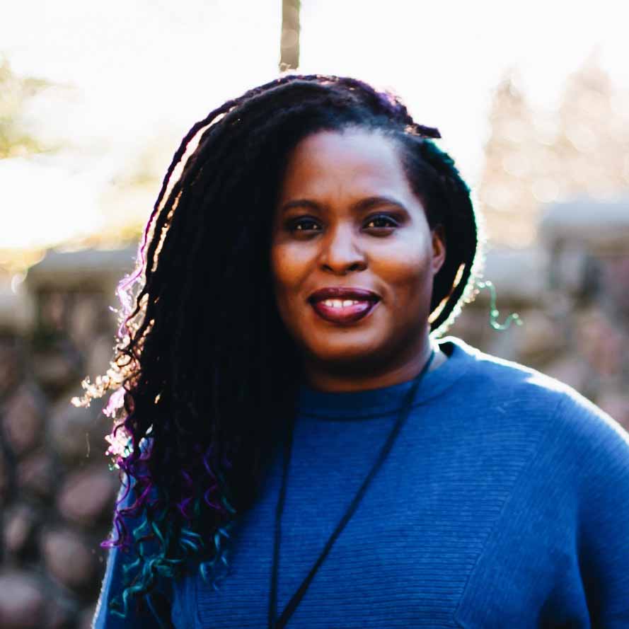 Smiling woman with long, side-swept hair with teal and purple tips, wearing a blue sweater, standing outdoors in soft sunlight.