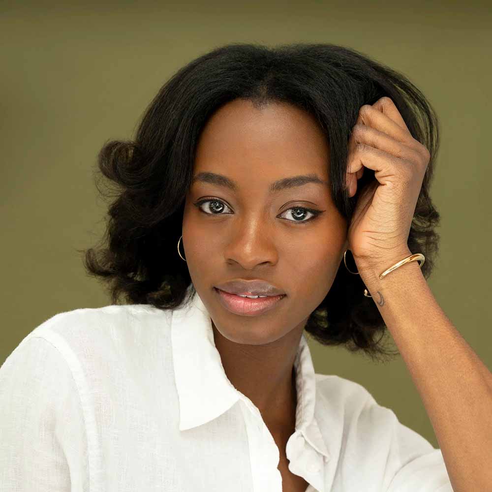Studio portrait of a woman with shoulder-length wavy hair wearing a white collared shirt and hoop earrings, resting their head on one hand against a muted green background.